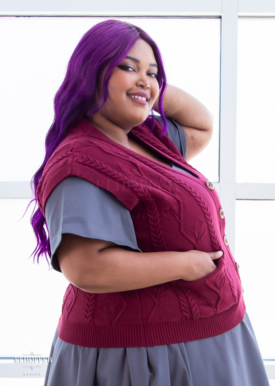 A side view of Jade, a light brown skinned 2xl model with long wavy purple hair, smiling while wearing the L (3XL-4XL) sample of a cranberry red button up knit vest with a leafy vine and cable knit pattern, light brown buttons, and front pockets over a charcoal grey knee length dress with short flutter sleeves.
