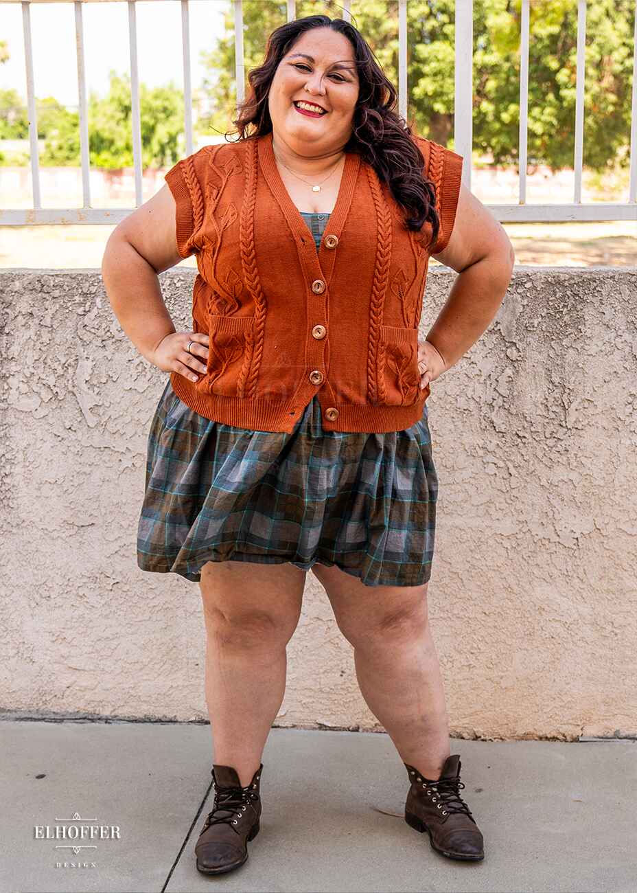 Alysia, a sun kissed skin 2xl model with long dark brown curly hair, is smilling while wearing a pumpkin orange button up knit vest with a leafy vine and cable knit pattern, light brown buttons, and front pockets.