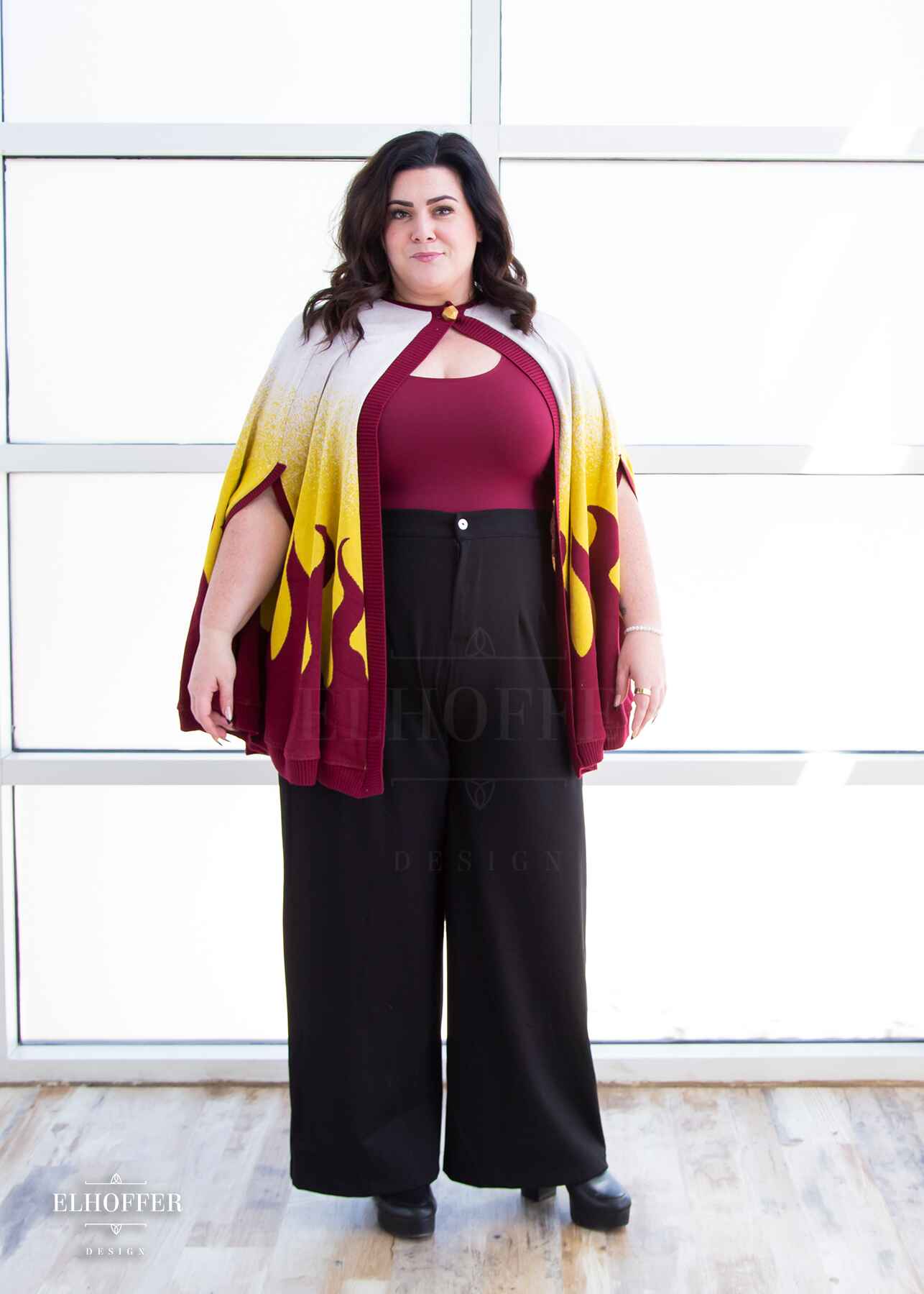 Stacy, a light skinned XL model with shoulder length wavy dark brown hair, is wearing a below hip length knit cape. The cape is a gradient of color from white at the shoulders, to yellow, to a red fire design at the bottom, it also has a button closer at the neck, and armholes in the side seam. She paired the cape with a burgundy scoop neck crop top and black pleated trousers.