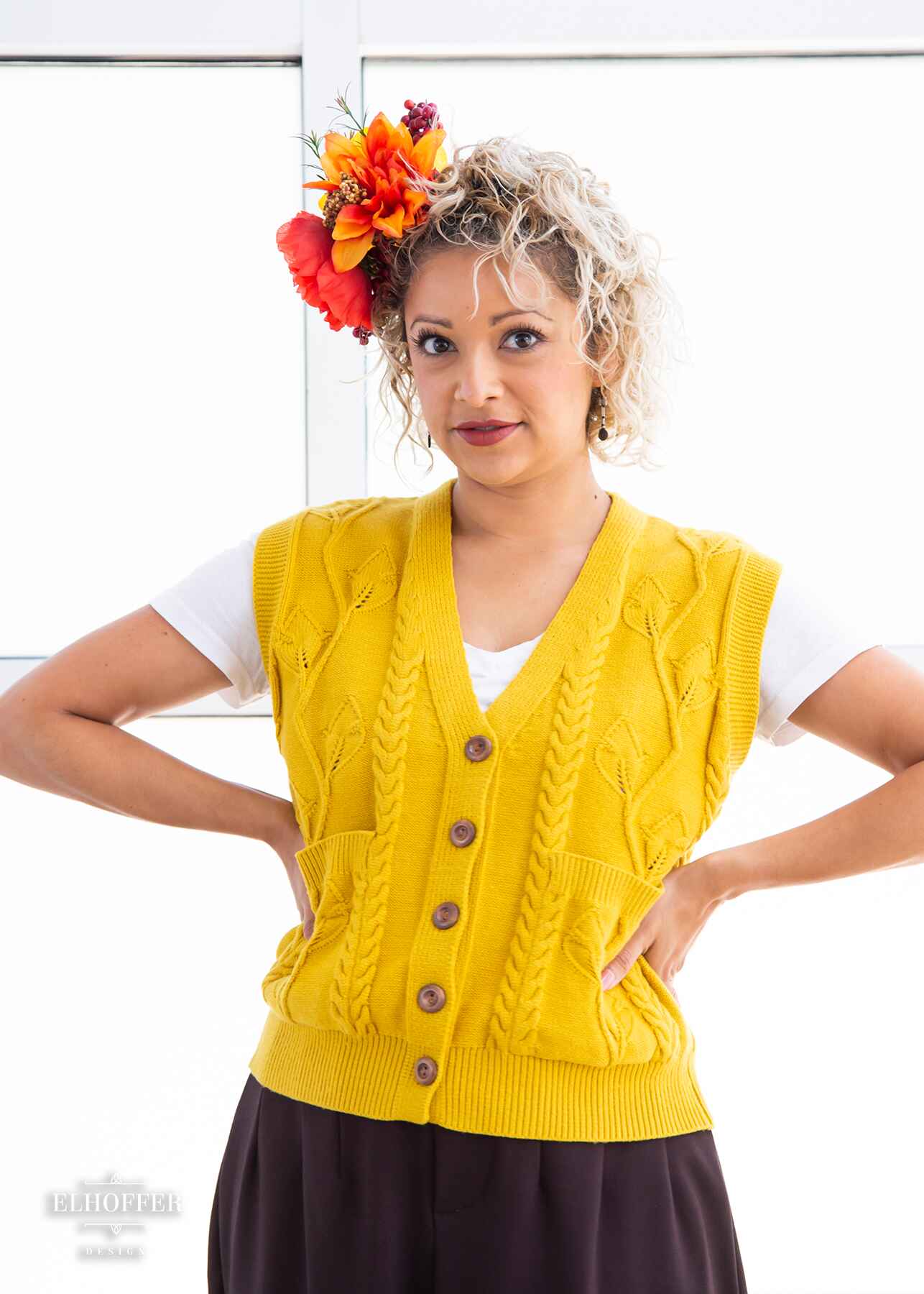 Simone, a light olive skinned S model with short platinum blonde curly hair, is wearing a golden yellow button up knit vest with a leafy vine and cable knit pattern, light brown buttons, and front pockets.