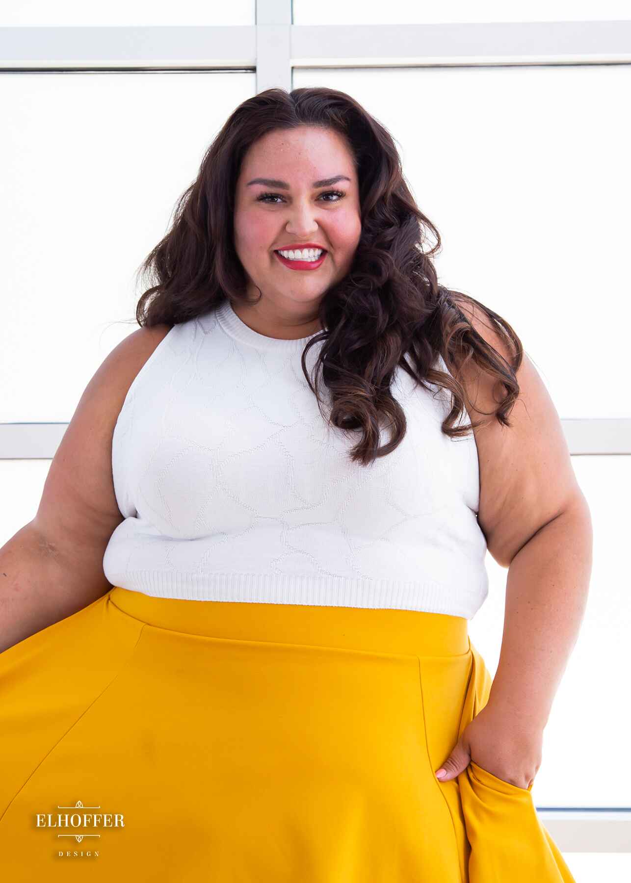 Kristen, an olive skinned 3xl model with long dark brown wavy hair, is smiling while wearing a bright white sleeveless knit crop top with a butterfly wing textured pattern. She paired the crop top with a golden yellow knee length high low skirt.