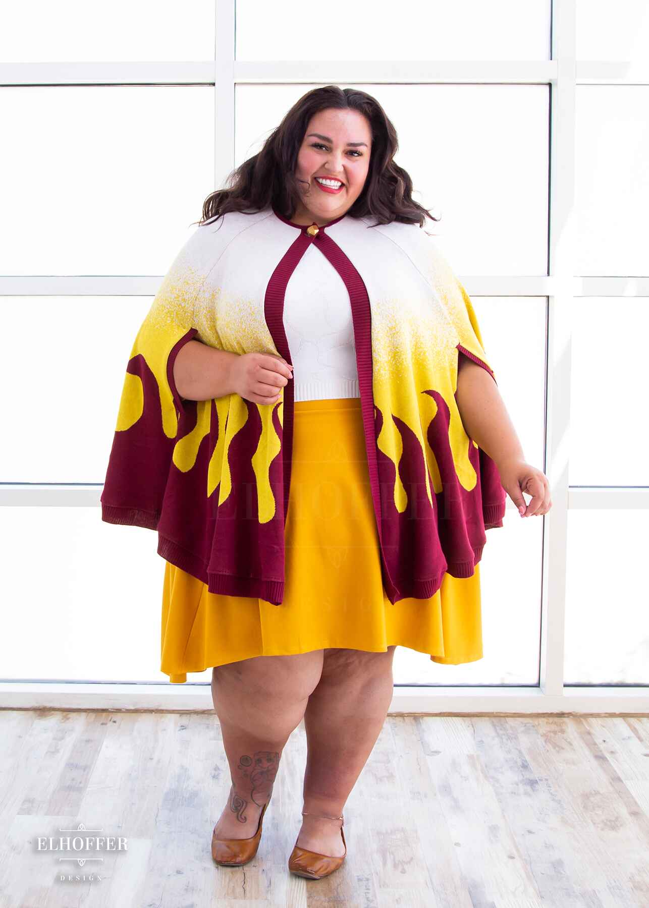 Kristen, a sun kissed skin 4xl model with shoulder length wavy dark brown hair, is smiling while wearing a below hip length knit cape. The cape is a gradient of color from white at the shoulders, to yellow, to a red fire design at the bottom, it also has a button closer at the neck, and armholes in the side seam. She paired the cape with a white knit crop top and a golden yellow knee length high low skirt.