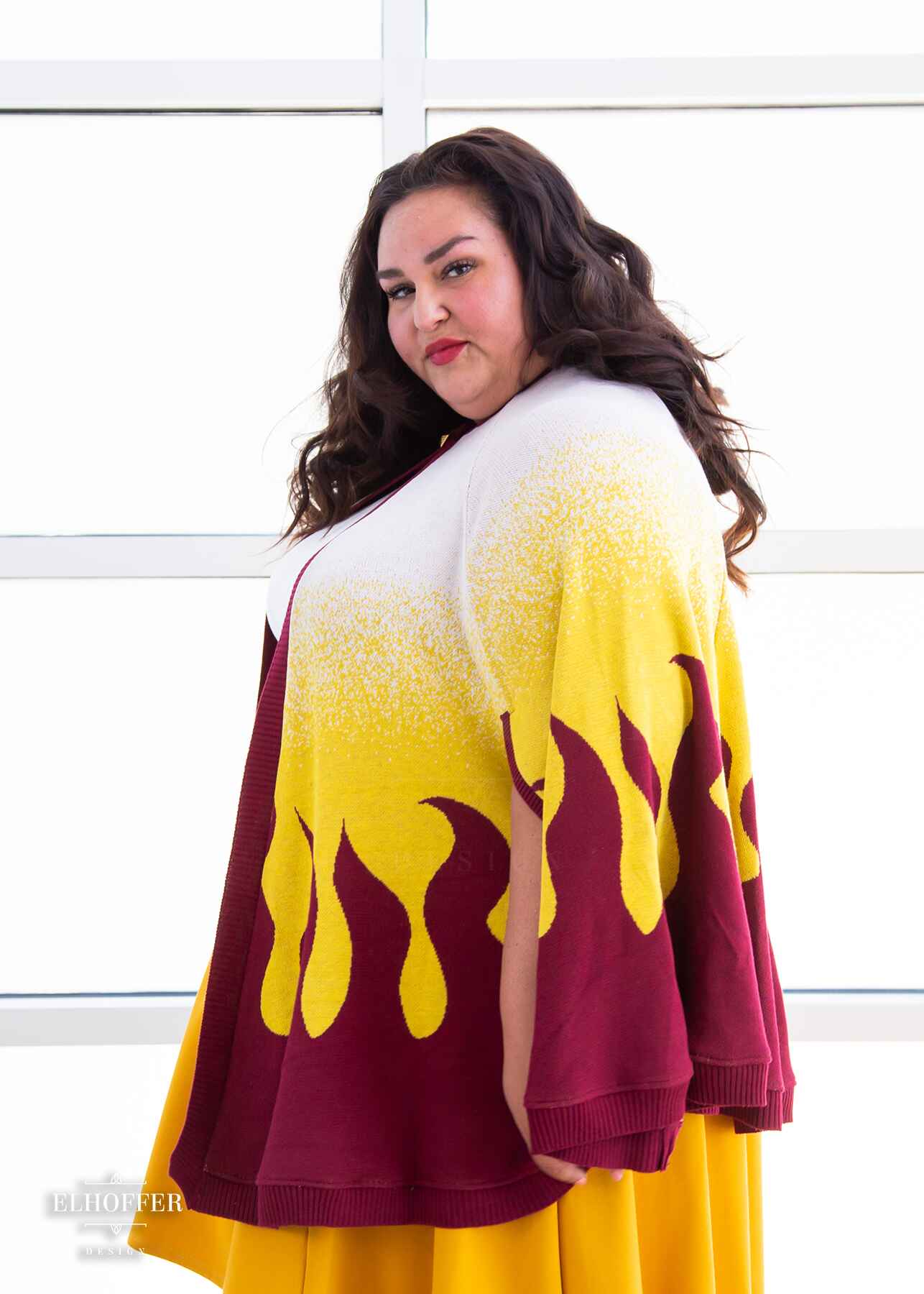 A side of Kristen, a sun kissed skin 4xl model with shoulder length wavy dark brown hair, wearing a below hip length knit cape. The cape is a gradient of color from white at the shoulders, to yellow, to a red fire design at the bottom, it also has a button closer at the neck, and armholes in the side seam. She paired the cape with a white knit crop top and a golden yellow knee length high low skirt.