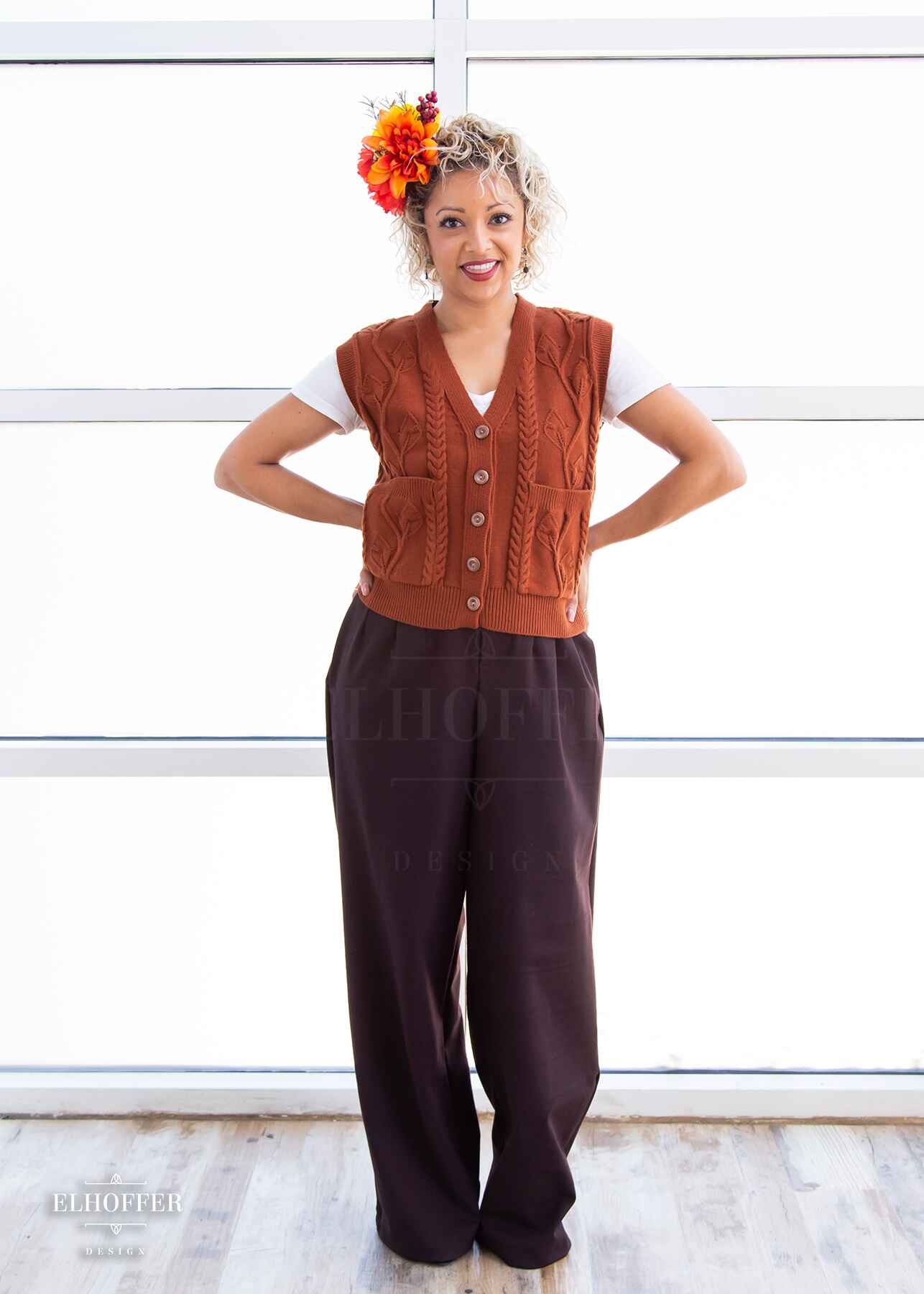 Simone, a light olive skinned S model with short curly platinum blonde hair, is smiling while wearing a pumpkin orange button up knit vest with a leafy vine and cable knit pattern, light brown buttons, and front pockets.