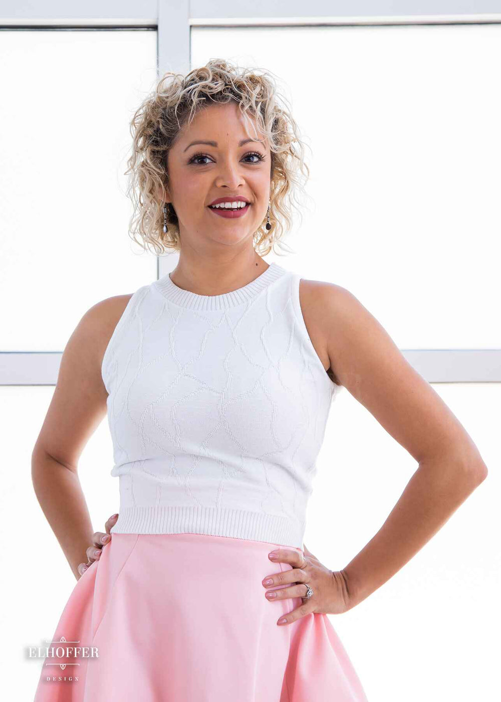 Simone, a light olive skinned S model with short curly platinum blonde hair, is wearing a bright white sleeveless knit crop top with a butterfly wing textured pattern. She paired the crop top with a light pink knee length high low skirt. 