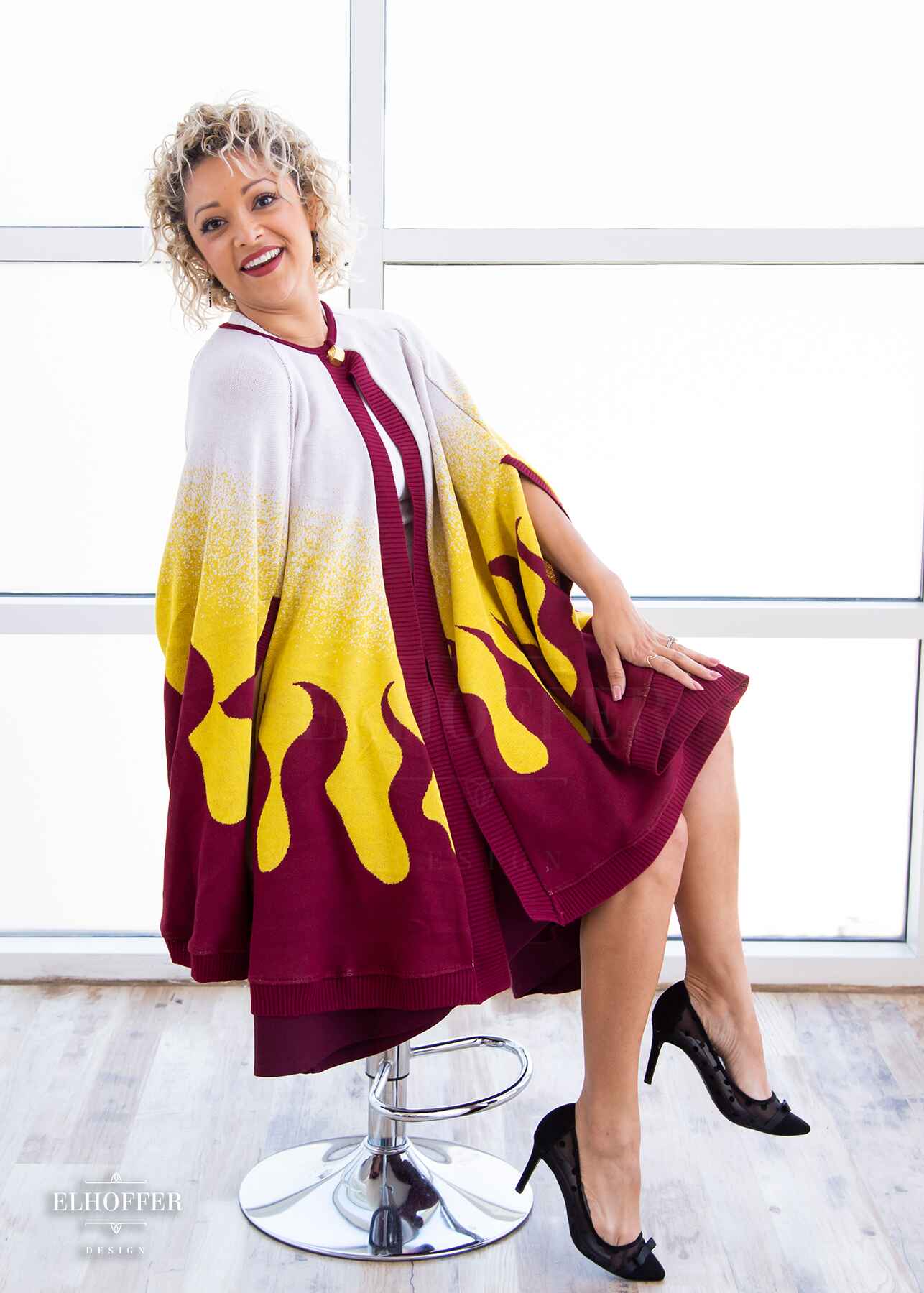 Simone, a size small model with light olive skin and curly platinum hair, is wearing a below hip length knit cape. The cape is a gradient of color from white at the shoulders, to yellow, to a red fire design at the bottom, it also has a button closer at the neck, and armholes in the side seam.