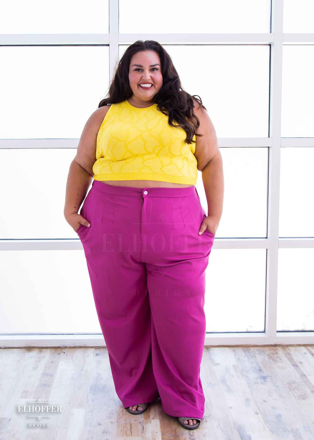 Kristen, an olive skinned 3xl model with long dark brown wavy hair, is smiling while wearing a bright yellow sleeveless knit crop top with a butterfly wing textured pattern.  She paired the crop top with pink trousers.