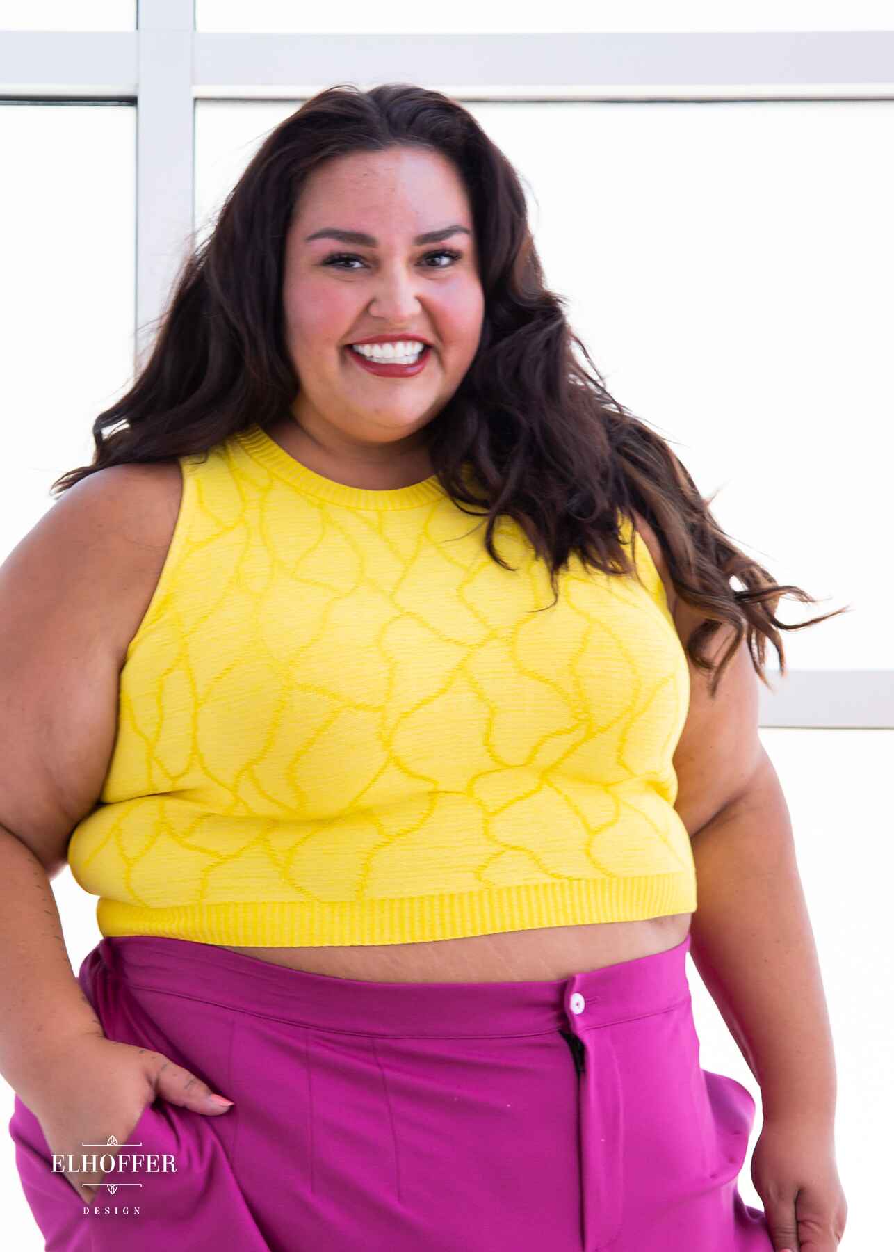 Kristen, an olive skinned 3xl model with long dark brown wavy hair, is smiling while wearing a bright yellow sleeveless knit crop top with a butterfly wing textured pattern.  She paired the crop top with pink trousers.