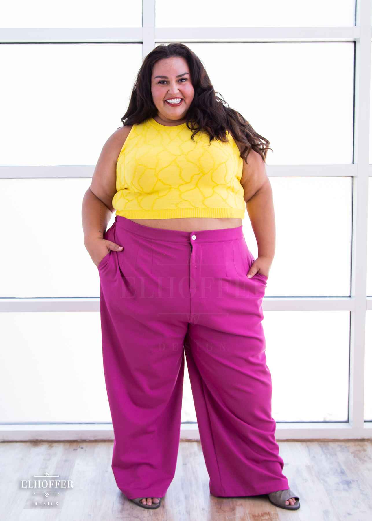 Kristen, an olive skinned 3xl model with long dark brown wavy hair, is smiling while wearing a bright yellow sleeveless knit crop top with a butterfly wing textured pattern.  She paired the crop top with pink trousers.