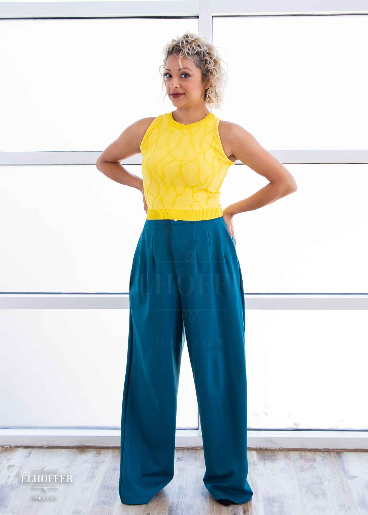 Simone, a light olive skinned S model with short curly platinum blonde hair, is wearing a bright yellow sleeveless knit crop top with a butterfly wing textured pattern. She paired the crop top with peacock teal trousers.