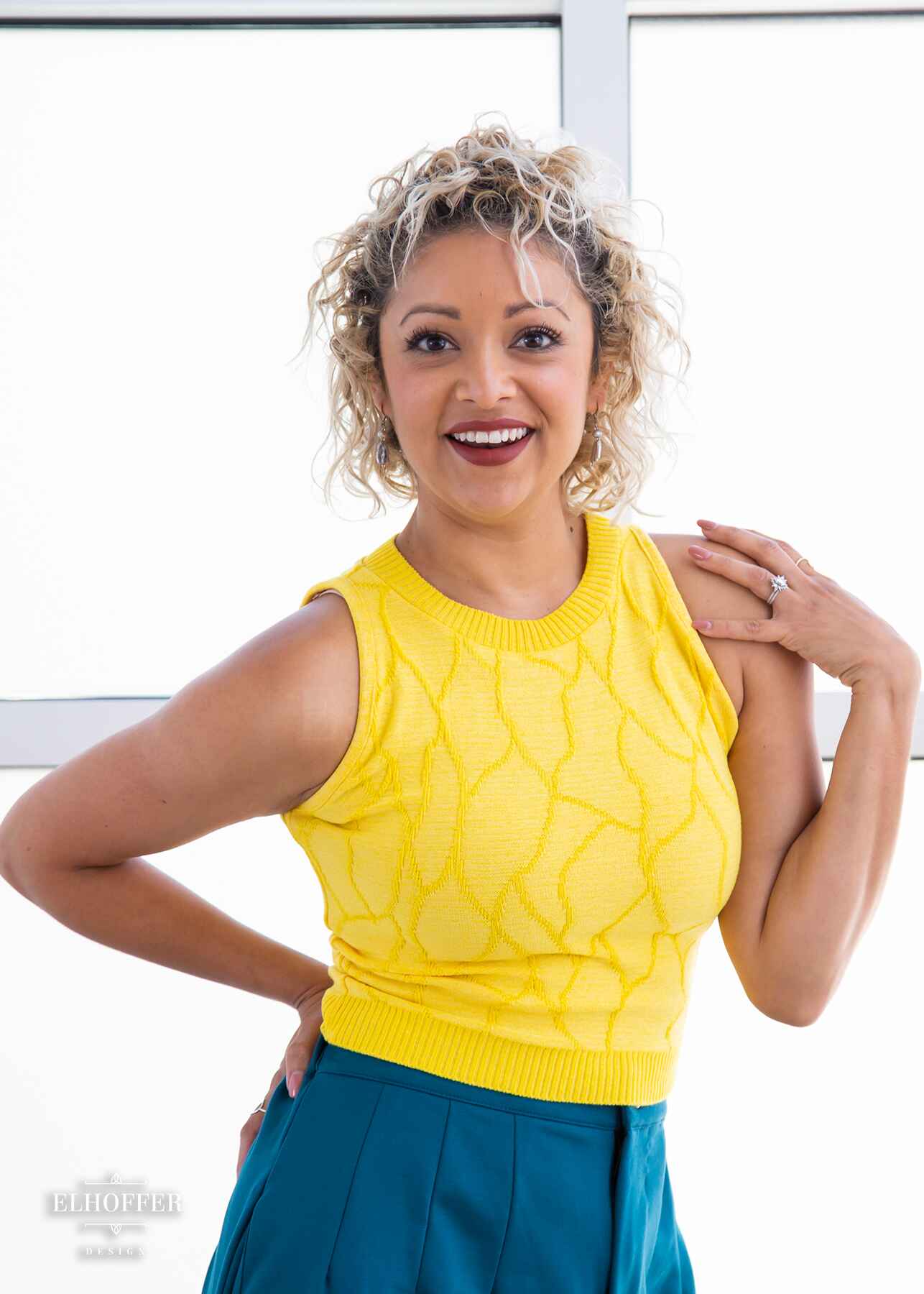 Simone, a light olive skinned S model with short curly platinum blonde hair, is smiling while wearing a bright yellow sleeveless knit crop top with a butterfly wing textured pattern. She paired the crop top with peacock teal trousers.
