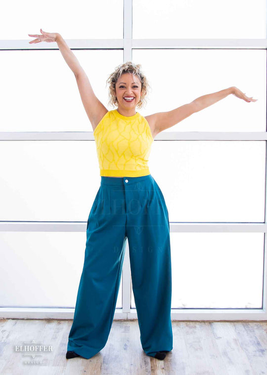 Simone, a light olive skinned S model with short curly platinum blonde hair, is smiling while wearing a bright yellow sleeveless knit crop top with a butterfly wing textured pattern. She paired the crop top with peacock teal trousers.