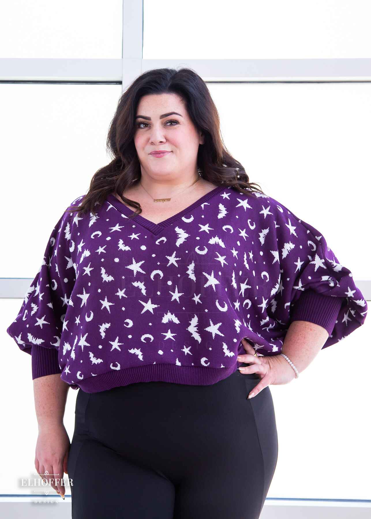 Stacy, a light skinned XL model with shoulder length wavy dark brown hair, is wearing an oversized v neck cropped sweater with batwing sleeves that gather at the wrist.  The main body of the sweater is purple with a white bat, star, moon, and dot pattern repeated throughout.