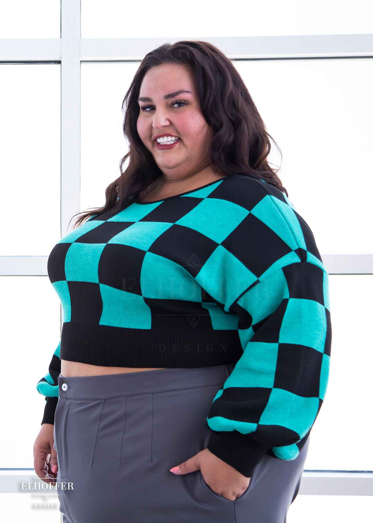 Kristen, a sun kissed skin 3XL model with long dark brown hair, is smiling while wearing a cropped oversize sweater with a black and green chessboard pattern and long billowing sleeves.  She paired the sweater with medium grey wide leg slacks.