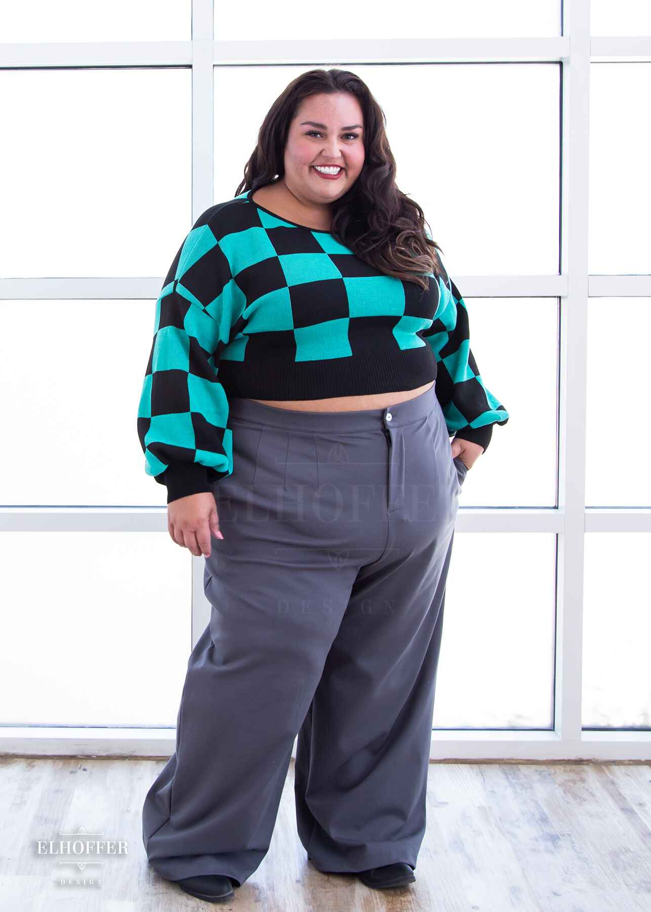 Kristen, a sun kissed skin 3XL model with long dark brown hair, is smiling while wearing a cropped oversize sweater with a black and green chessboard pattern and long billowing sleeves.  She paired the sweater with medium grey wide leg slacks.