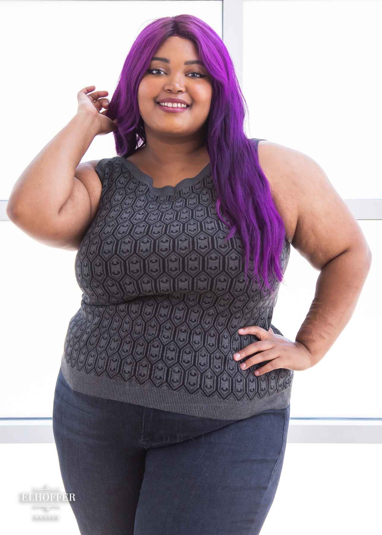 Jade, a light brown skinned 2xl model with long wavy purple hair, is smiling while wearing a knit sleeveless top with a boatneck and an armor plated design