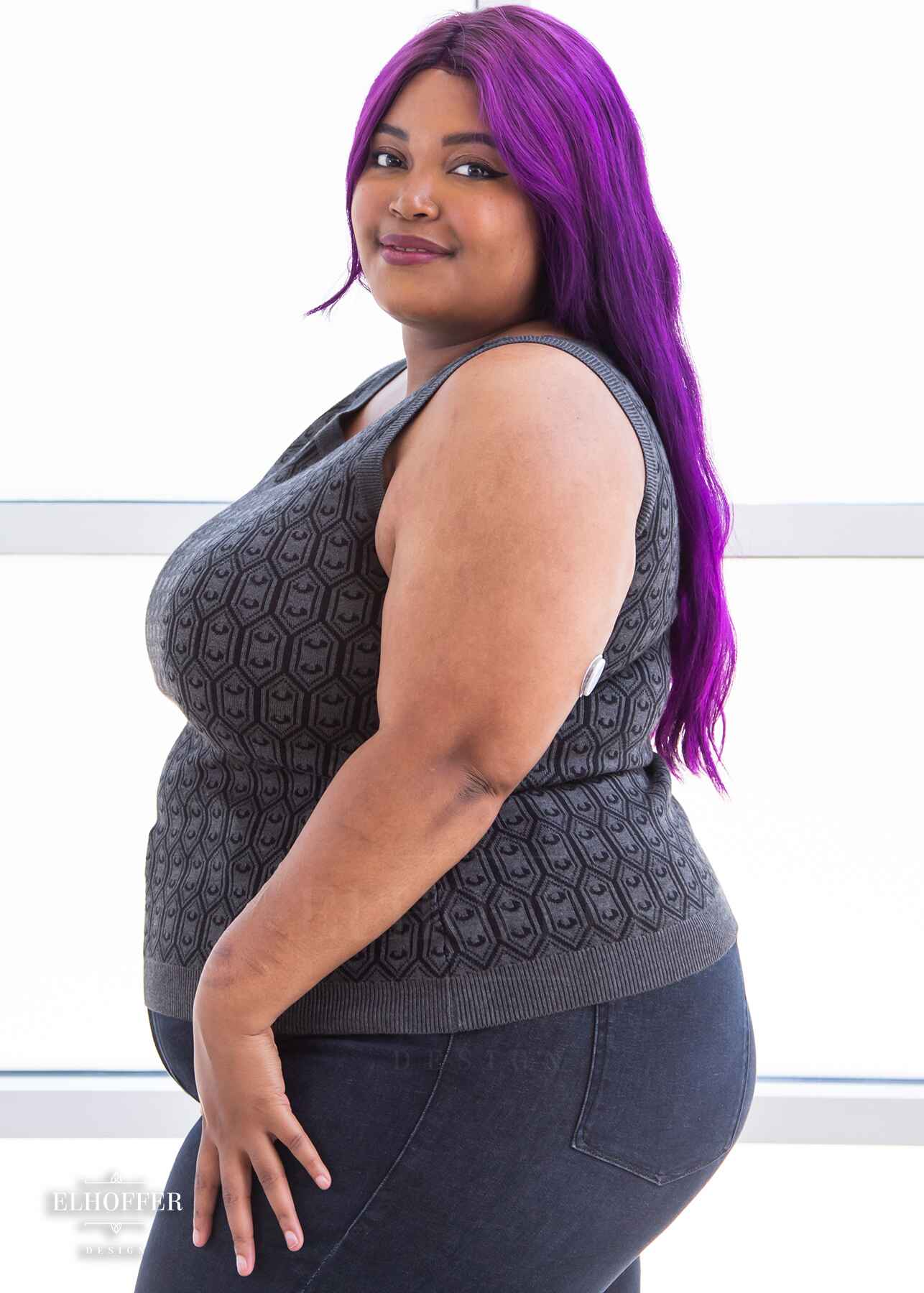 A side view of Jade, a light brown skinned 2xl model with long wavy purple hair, wearing a knit sleeveless top with a boatneck and an armor plated design