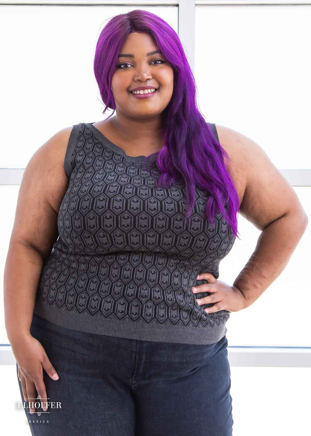 Jade, a light brown skinned 2xl model with long wavy purple hair, is smiling while wearing a knit sleeveless top with a boatneck and an armor plated design