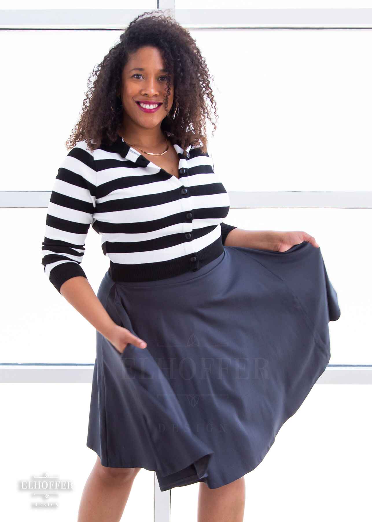 Francesca, a light skinned L model with dark brown shoulder length tight curly hair, is wearing a cropped knit button up cardigan with long sleeves, black and white horizontal stripes, and a black hood.
