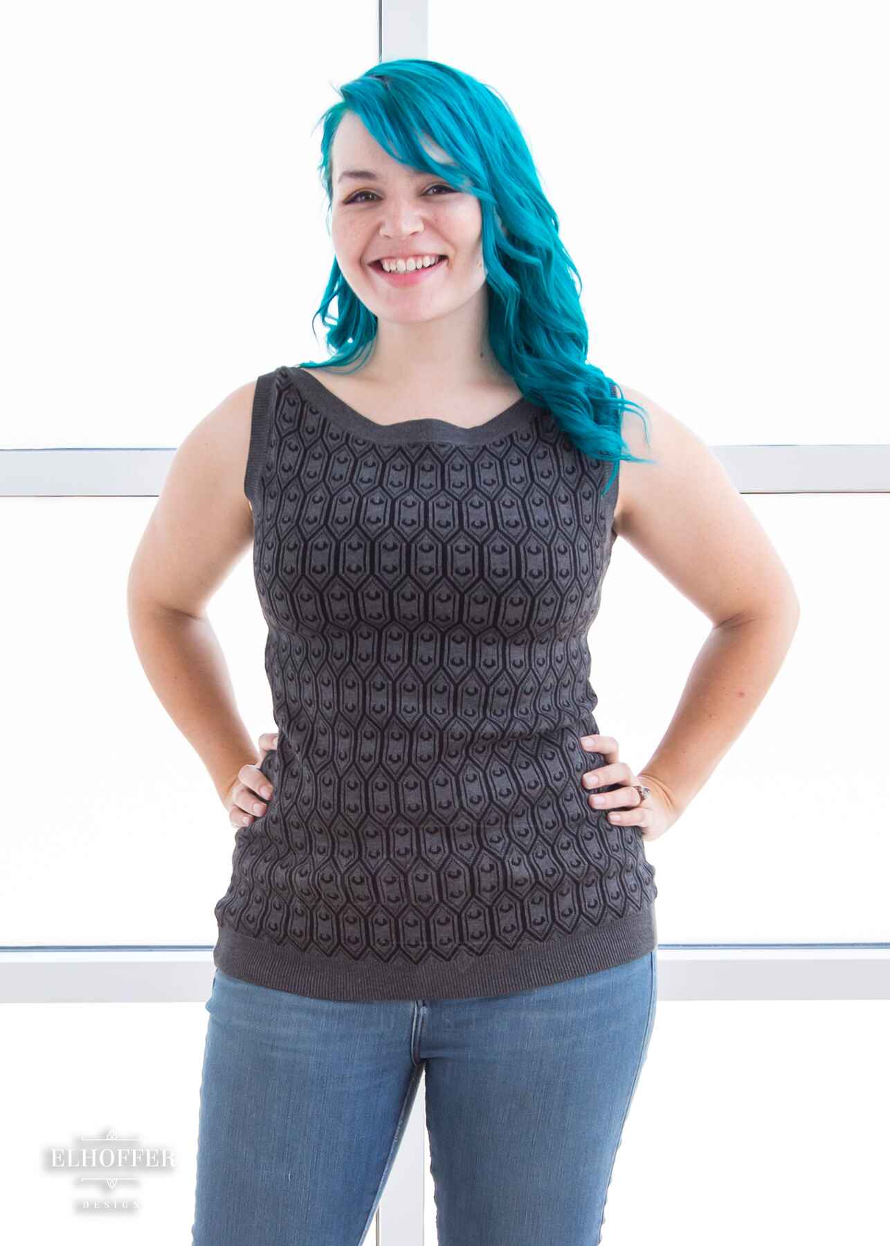 Kiri, a fair skinned S model with long wavy teal hair, is smiling while wearing a knit sleeveless top with a boatneck and an armor plated design.