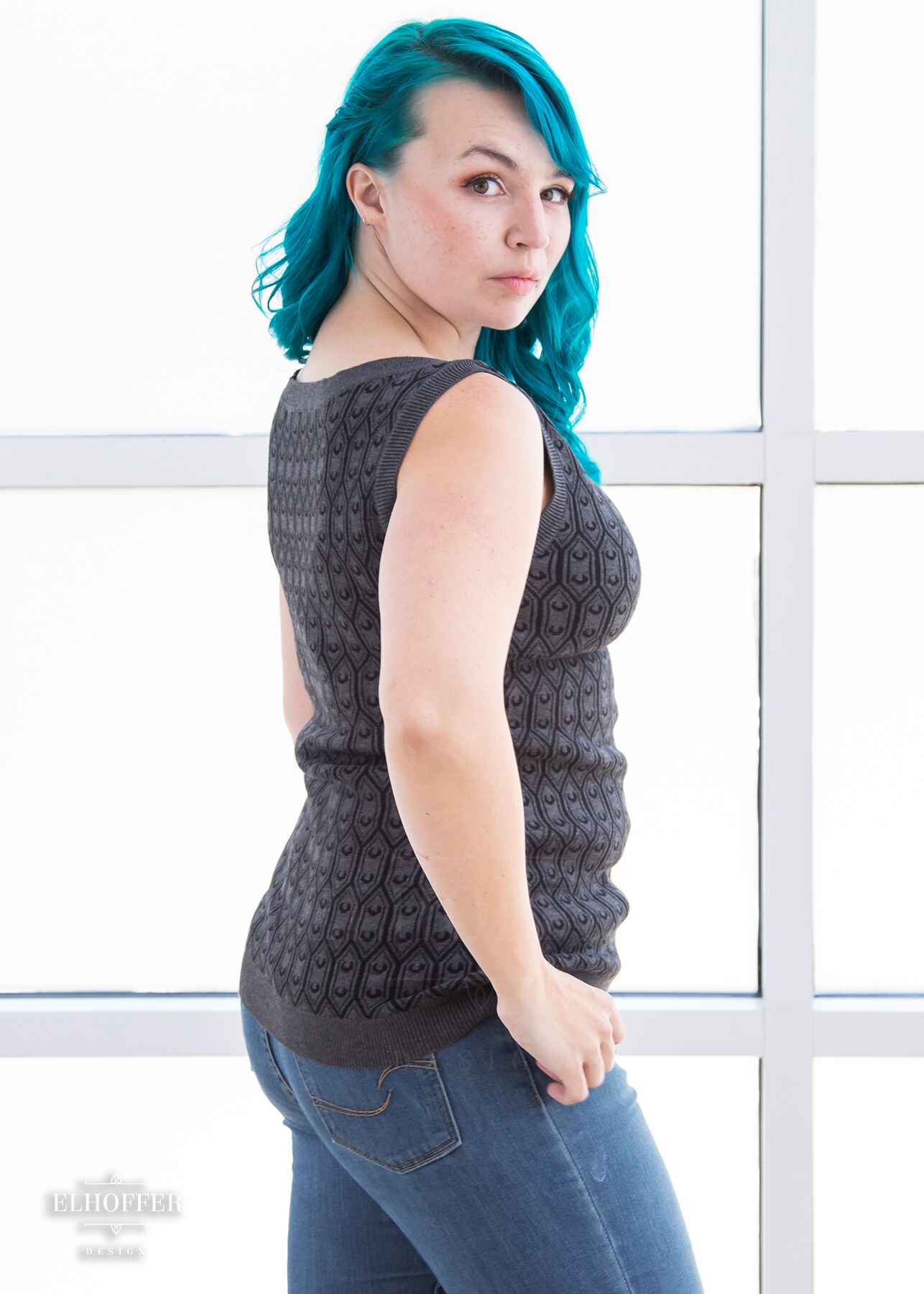 A side view of Kiri, a fair skinned S model with long wavy teal hair, wearing a knit sleeveless top with a boatneck and an armor plated design.