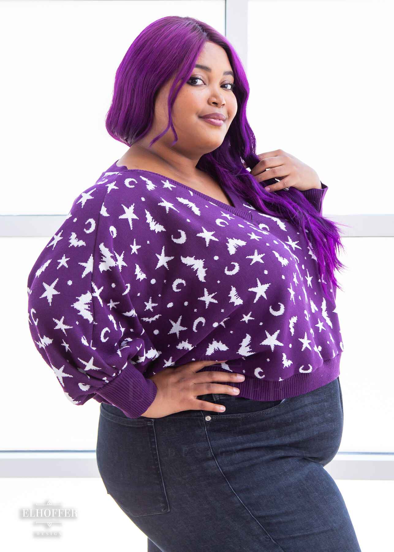A side view of Jade, a light skinned 2xl model with long wavy purple hair, wearing an oversized v neck cropped sweater with batwing sleeves that gather at the wrist. The main body of the sweater is purple with a white bat, star, moon, and dot pattern repeated throughout.