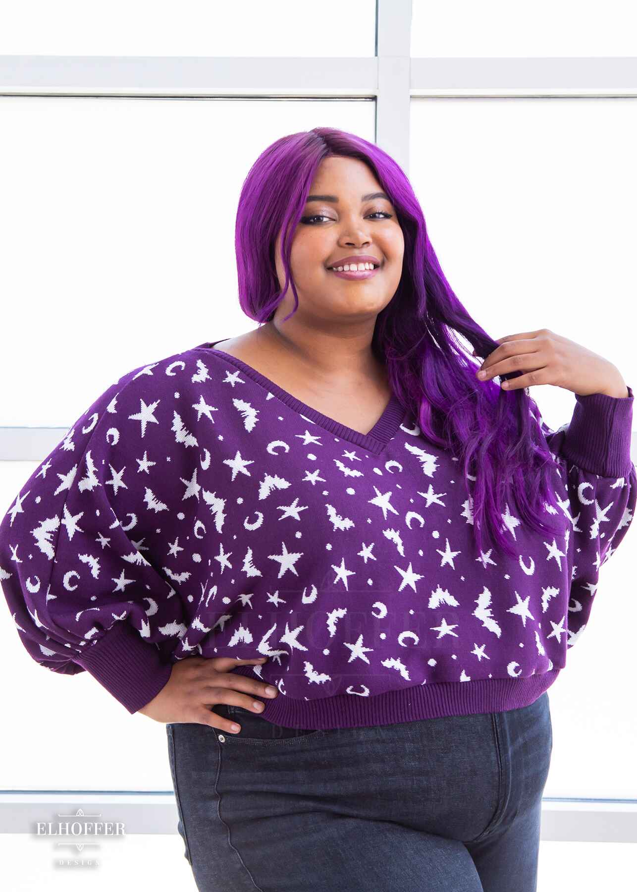 Jade, a light skinned 2xl model with long wavy purple hair, is smiling while wearing an oversized v neck cropped sweater with batwing sleeves that gather at the wrist. The main body of the sweater is purple with a white bat, star, moon, and dot pattern repeated throughout.