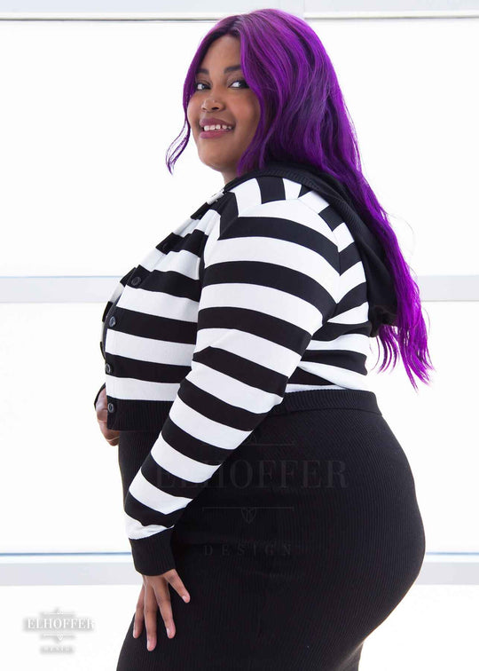 A side view of Jade, a medium dark skinned 2xl model with long wavy purple hair, smiling whlie wearing a cropped knit button up cardigan with long sleeves, black and white horizontal stripes, and a black hood.