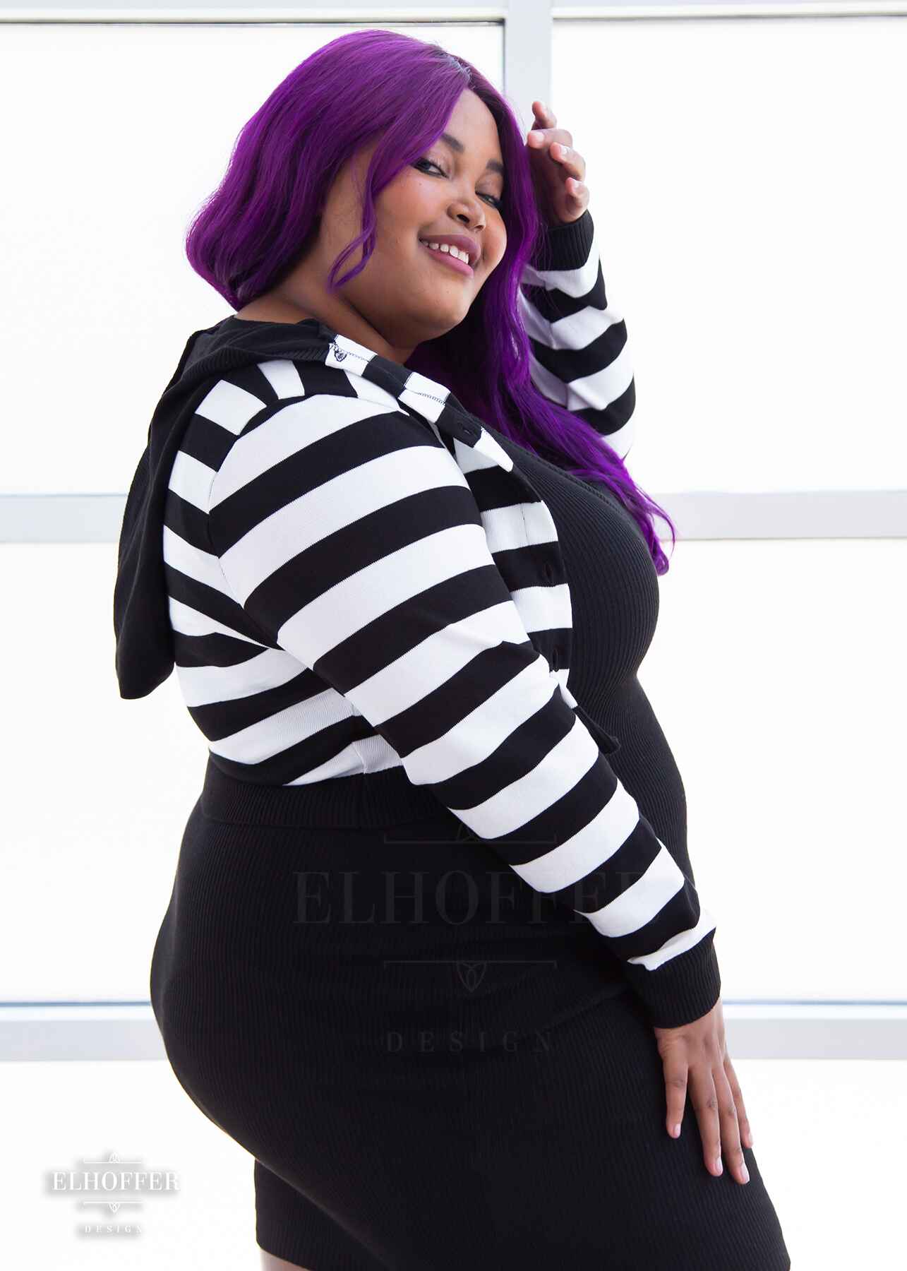A side view of Jade, a medium dark skinned 2xl model with long wavy purple hair, smiling whlie wearing a cropped knit button up cardigan with long sleeves, black and white horizontal stripes, and a black hood.