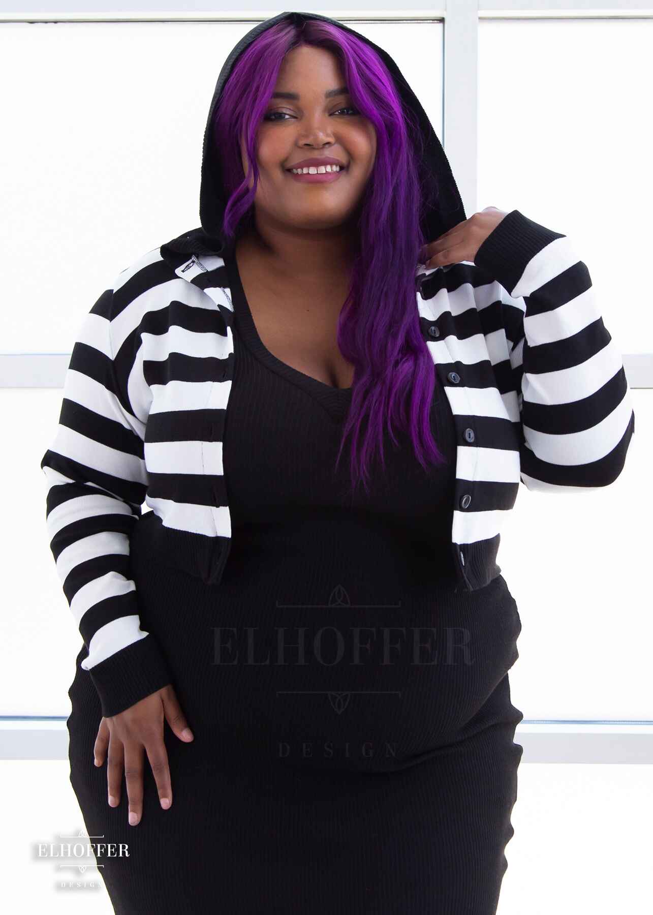 Jade, a medium dark skinned 2xl model with long wavy purple hair, is smiling whlie wearing a cropped knit button up cardigan with long sleeves, black and white horizontal stripes, and a black hood.