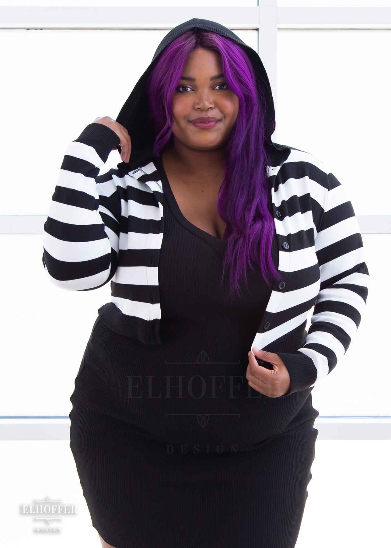 Jade, a medium dark skinned 2xl model with long wavy purple hair, is wearing a cropped knit button up cardigan with long sleeves, black and white horizontal stripes, and a black hood.