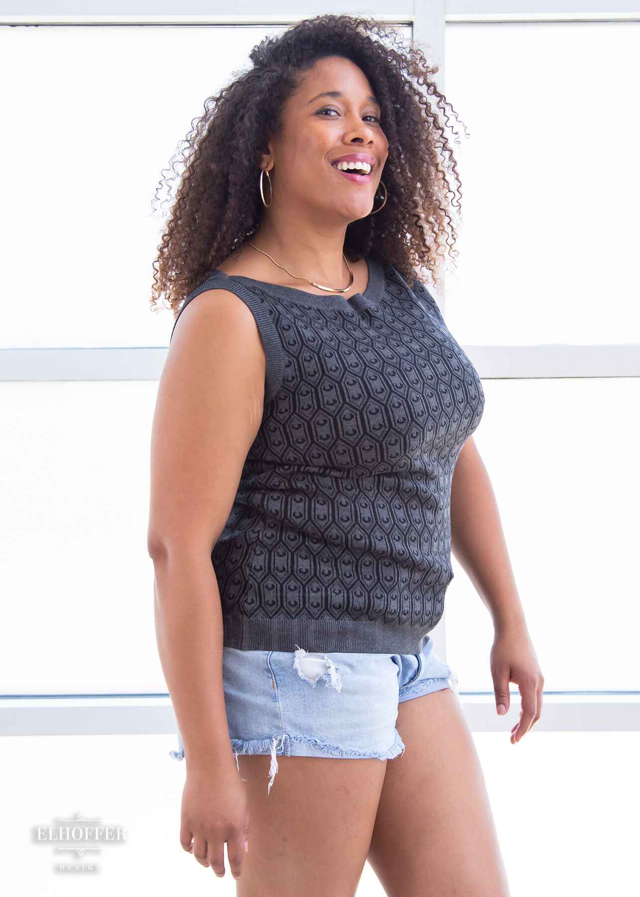 Francesca, a light brown skinned L model with long dark brown tight curly hair, is smiling while wearing a knit sleeveless top with a boatneck and an armor plated design.