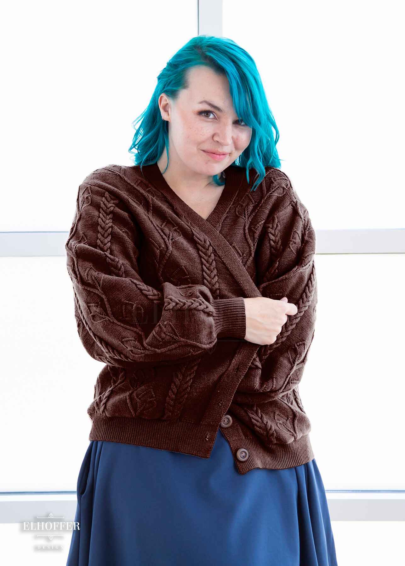 Kiri, a fair skinned S model with long wavy teal hair, is wearing the S (XS-M) sample of a chocolate brown knit long sleeve button up cardigan with a leafy vine and cable knit pattern, long billowing sleeves, light brown buttons, and front pockets over a denim blue knee length dress.