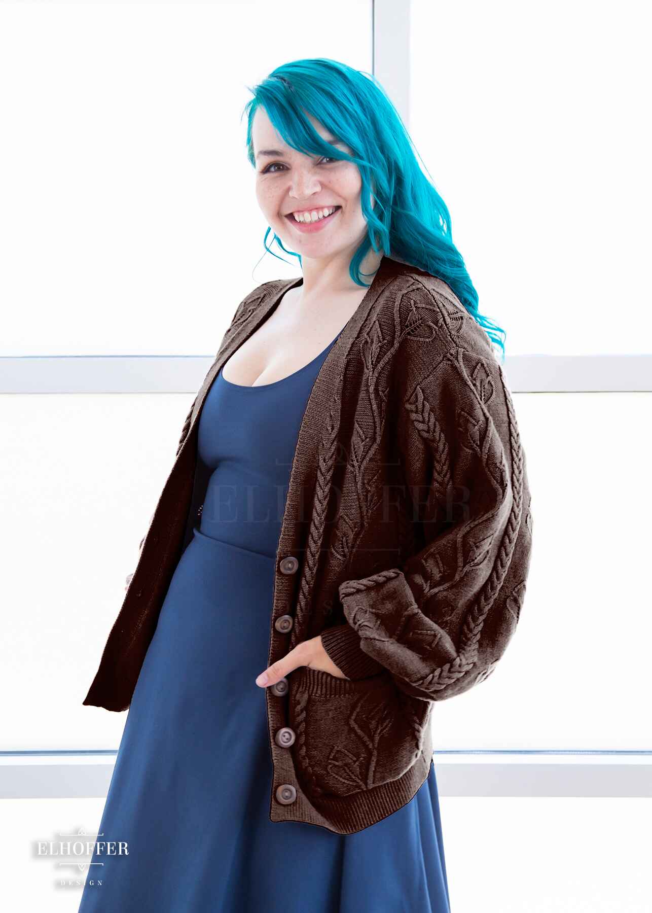 Kiri, a fair skinned S model with long wavy teal hair, is smiling while wearing the S (XS-M) sample of a chocolate brown knit long sleeve button up cardigan with a leafy vine and cable knit pattern, long billowing sleeves, light brown buttons, and front pockets over a denim blue knee length dress.