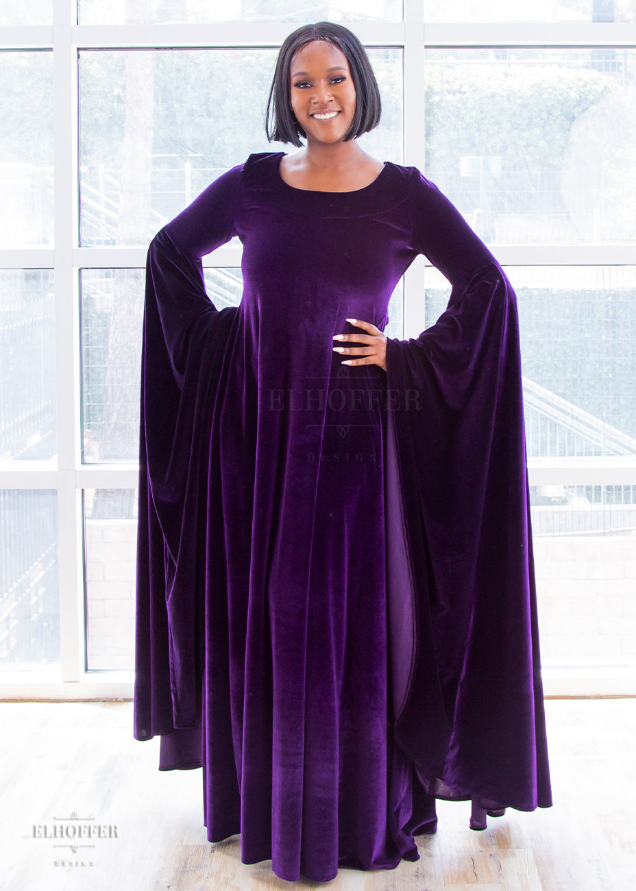 Lynsi (a medium dark skinned size large model with short dark hair) wears the purple velvet floor length dress. It has a boatneck neckline and extra long flowing sleeves that fall to ankle length.