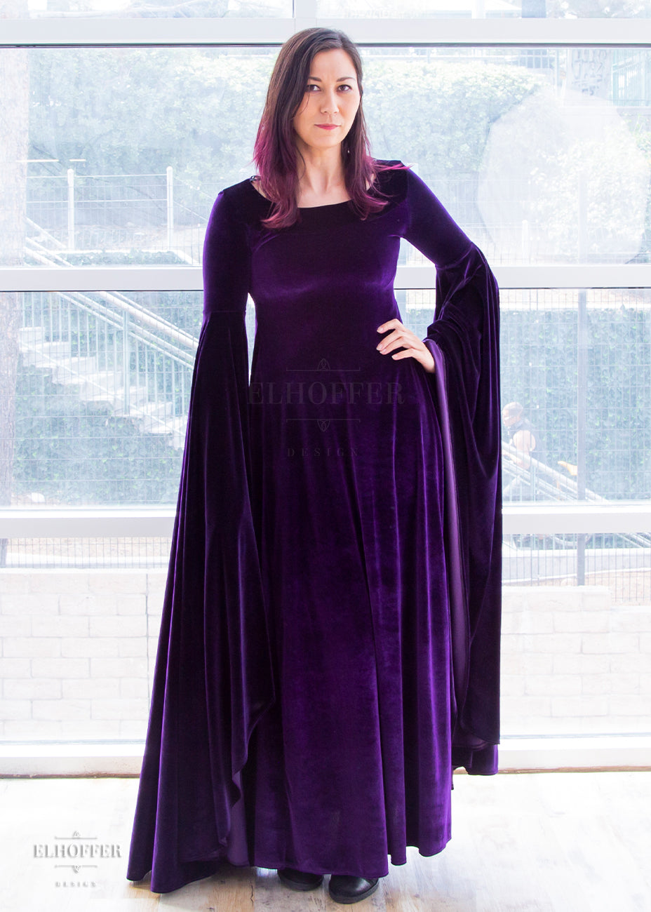 Susan (a medium fair size extra small model with dark hair with a purple ombre) wears the purple velvet floor length dress. It has a boatneck neckline and extra long flowing sleeves that fall to ankle length.