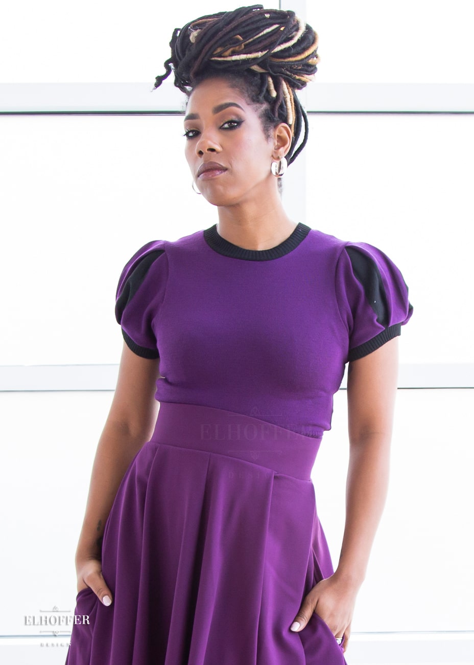 Krystina, a medium dark skinned S model with long braids in an updo, is wearing a purple knit short sleeve crop top with black ribbing along neckline and cuffs.  The sleeves are split puff sleeves with alternating colors of purple and black. She paired the knit top with a plum skater skirt.