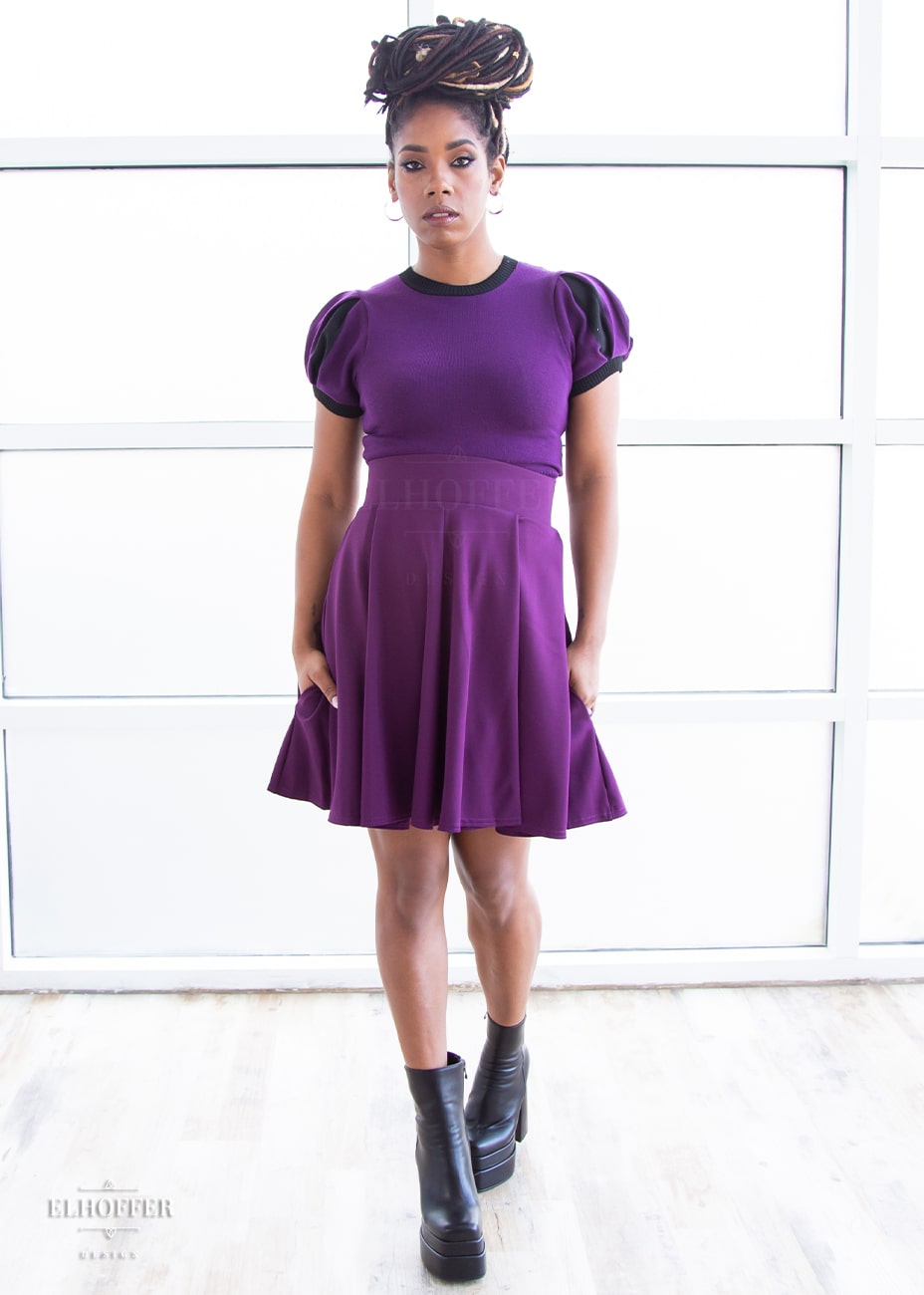 Krystina, a medium dark skinned S model with long braids in an updo, is wearing a purple knit short sleeve crop top with black ribbing along neckline and cuffs.  The sleeves are split puff sleeves with alternating colors of purple and black. She paired the knit top with a plum skater skirt.