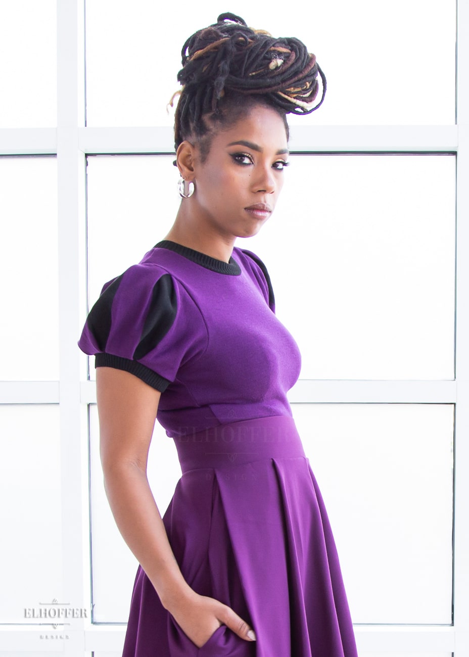 A side view of Krystina, a medium dark skinned S model with long braids in an updo, wearing a purple knit short sleeve crop top with black ribbing along neckline and cuffs.  The sleeves are split puff sleeves with alternating colors of purple and black. She paired the knit top with a plum skater skirt.