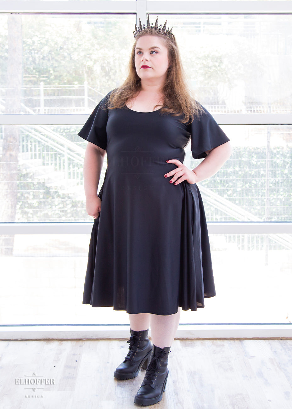 Lucy, a size large fair skinned model with long dark brown hair, is wearing a knee-length pullover dress with scoop neck, flutter sleeves, and side pockets in black.