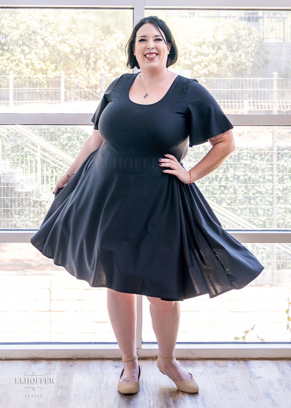 Katie Lynn, a size 2XL fair skinned model with short dark hair, is wearing a knee-length pullover dress with scoop neck, flutter sleeves, and side pockets in black.
