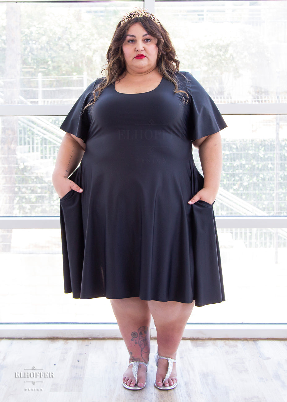 Kristen, a size 3XL olive skinned model with long brown highlighted hair, is wearing a knee-length pullover dress with scoop neck, flutter sleeves, and side pockets in black.