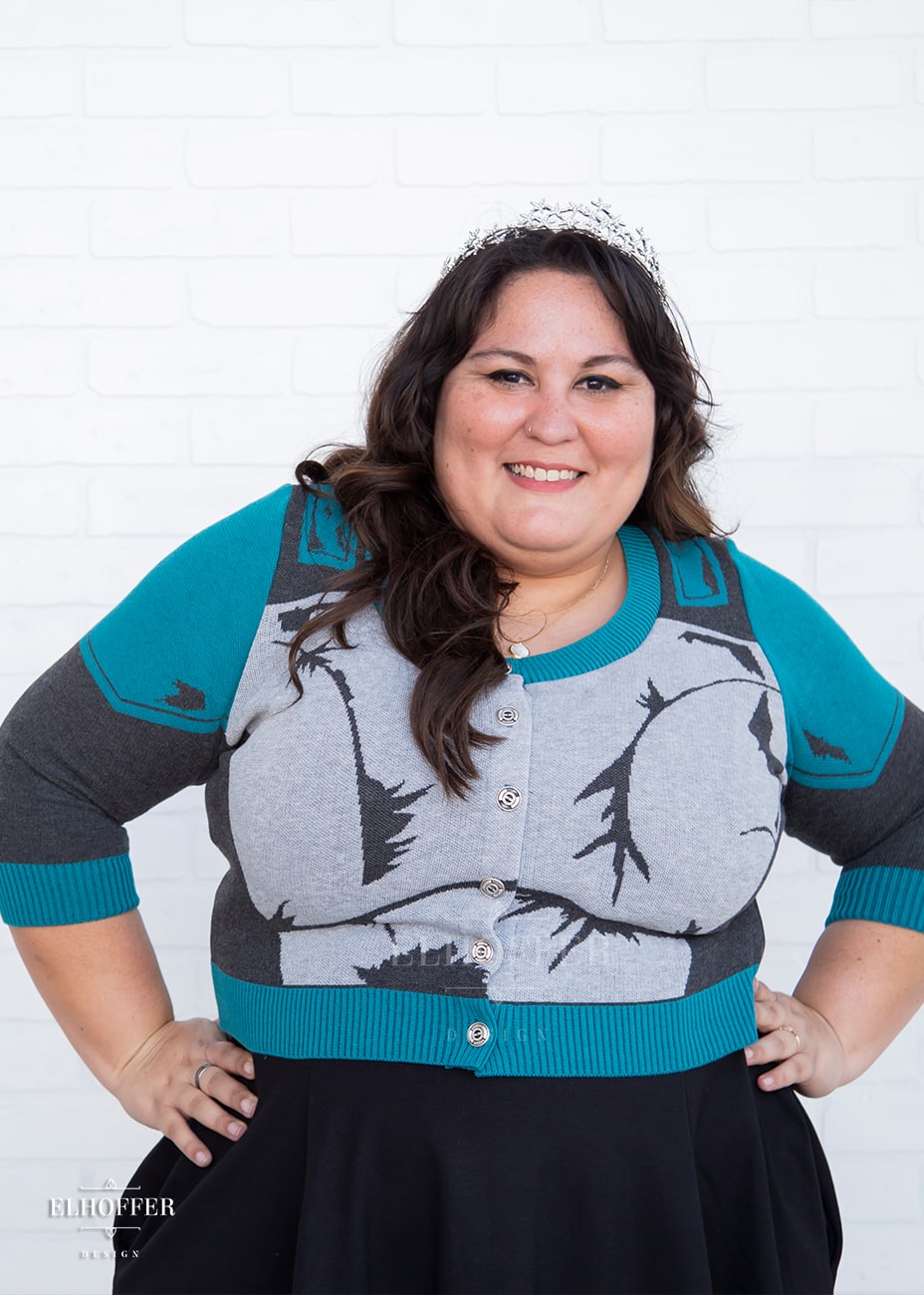 Alysia, a sun kissed skin 2xl model with long wavy dark brown hair, is smiling while wearing a cropped button up knit cardigan with 3/4 sleeves. The cardigan front is mainly a light grey with dark grey decorative lines that give the impression of battle damaged armor, with teal at the shoulders, and the bottom half of the sleeve and back of the cardigan are a dark grey. The neckine, bottom, and cuffs are all teal ribbing.