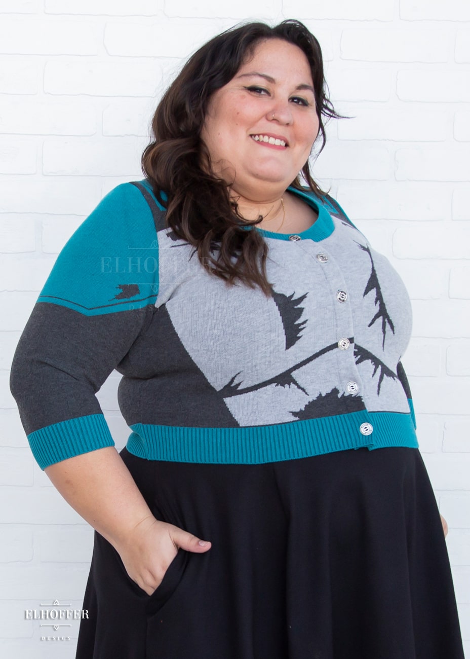 Alysia, a sun kissed skin 2xl model with long wavy dark brown hair, is smiling while wearing a cropped button up knit cardigan with 3/4 sleeves. The cardigan front is mainly a light grey with dark grey decorative lines that give the impression of battle damaged armor, with teal at the shoulders, and the bottom half of the sleeve and back of the cardigan are a dark grey. The neckine, bottom, and cuffs are all teal ribbing.