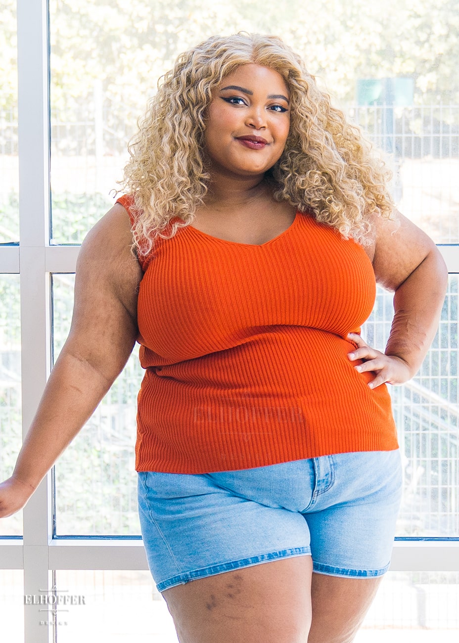 Jade, a medium dark skinned 2XL model with curly blonde hair,  is wearing a pullover sleeveless ribbed v-neck tank in a medium orange. The v-neck is actually reversible and offers two depths.