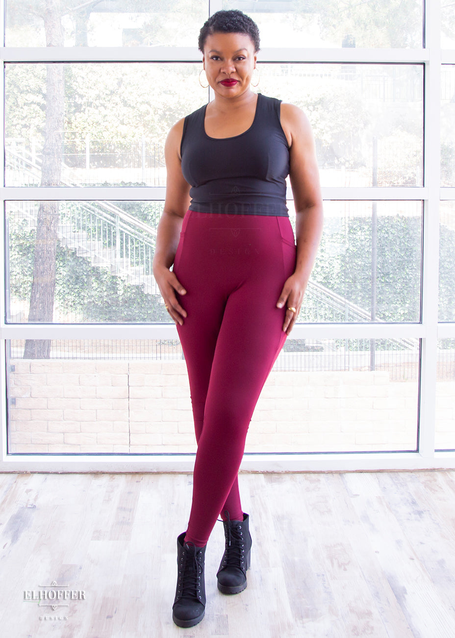 Desiree, a size large medium dark skinned model with very short dark hair, wearing a high-waisted full length fitted legging with side pockets built into the seamwork and a seamless front in a beautiful burgundy color. She is wearing them with a grey Jes crop.