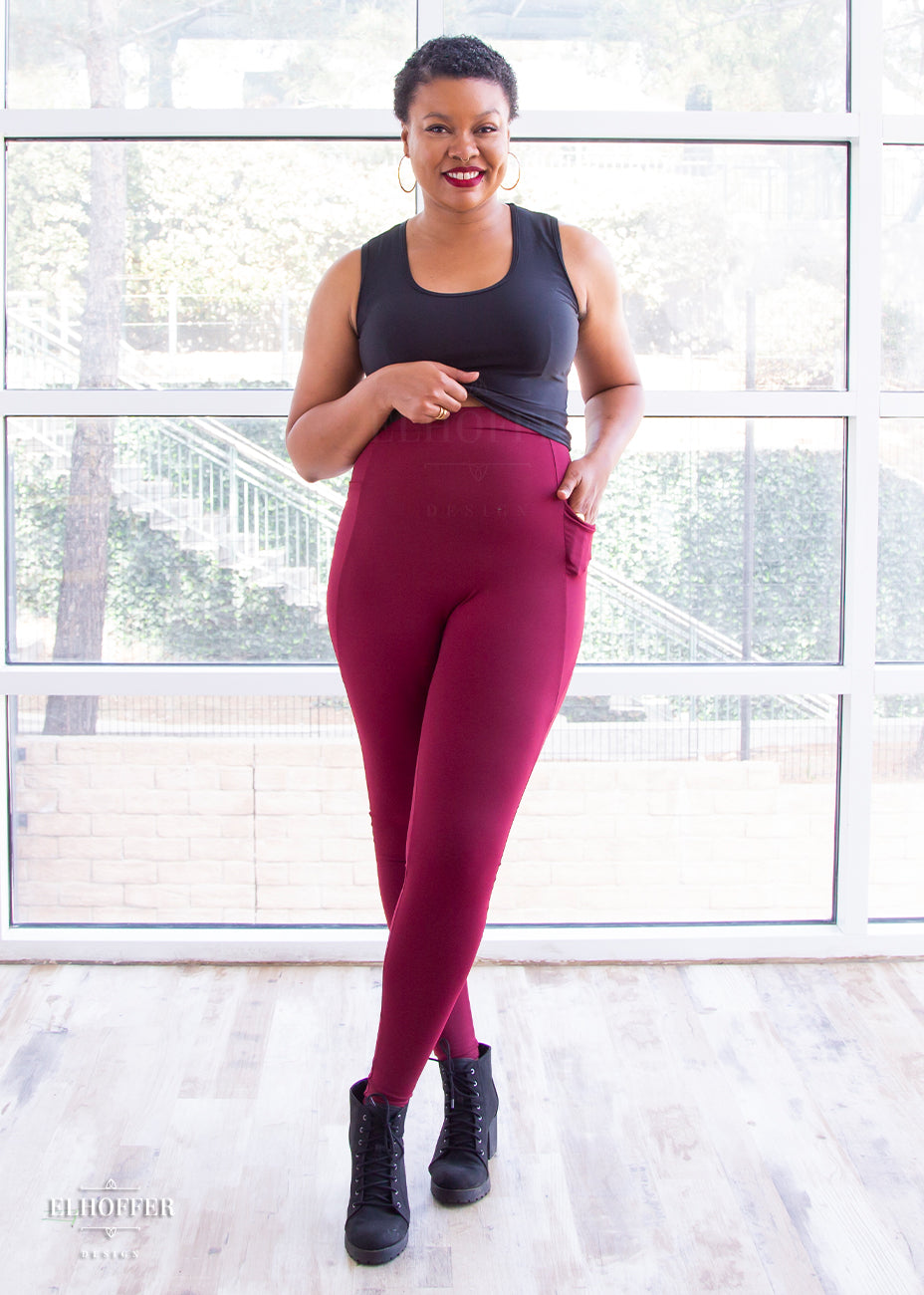 Desiree, a size large medium dark skinned model with very short dark hair, wearing a high-waisted full length fitted legging with side pockets built into the seamwork and a seamless front in a beautiful burgundy color. She is wearing them with a grey Jes crop. She lifts her crop to show off the high waist of the leggings.
