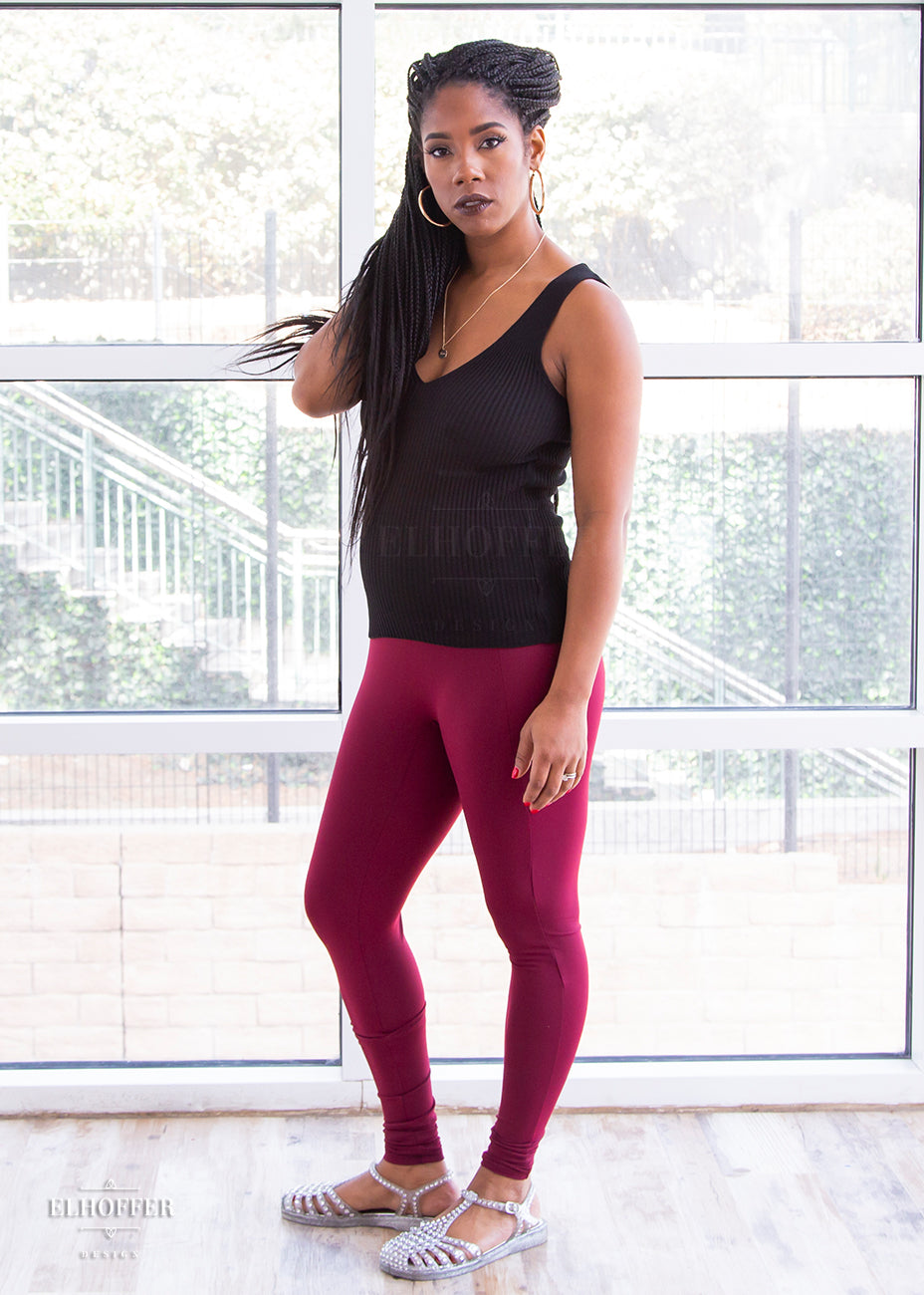 Krystina, a size small medium dark skinned model with long braids, wearing a high-waisted full length fitted legging with side pockets built into the seamwork and a seamless front in a beautiful burgundy color. She has paired them with a black knit Gemini top. 