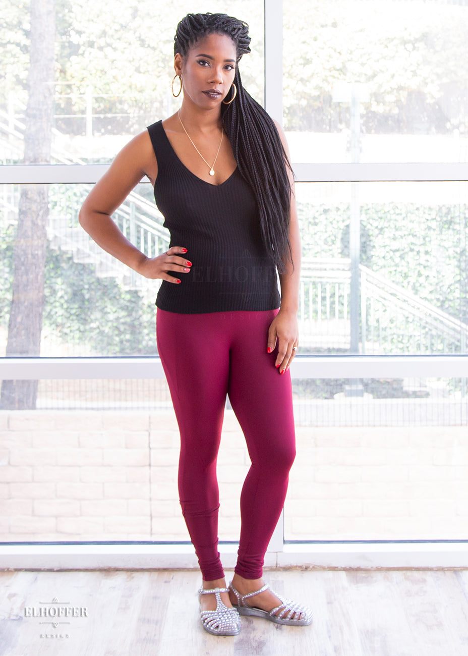 Krystina, a size small medium dark skinned model with long braids, wearing a high-waisted full length fitted legging with side pockets built into the seamwork and a seamless front in a beautiful burgundy color. She has paired them with a black knit Gemini top. 