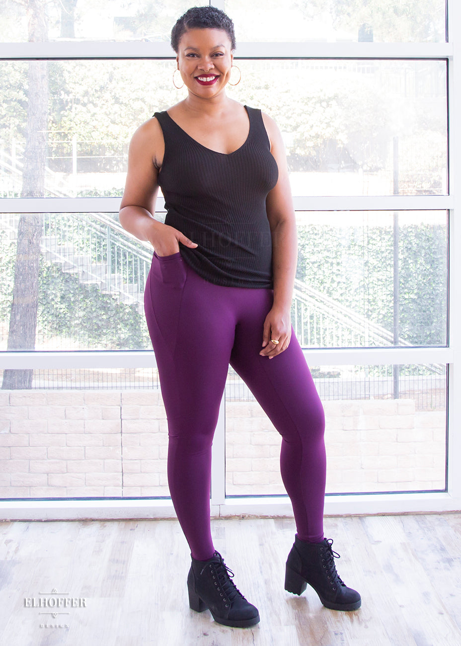 Desiree, a size large medium dark skinned model with very short dark hair, is wearing a pair of high waisted leggings with a seamless front and side seam pockets in plum.